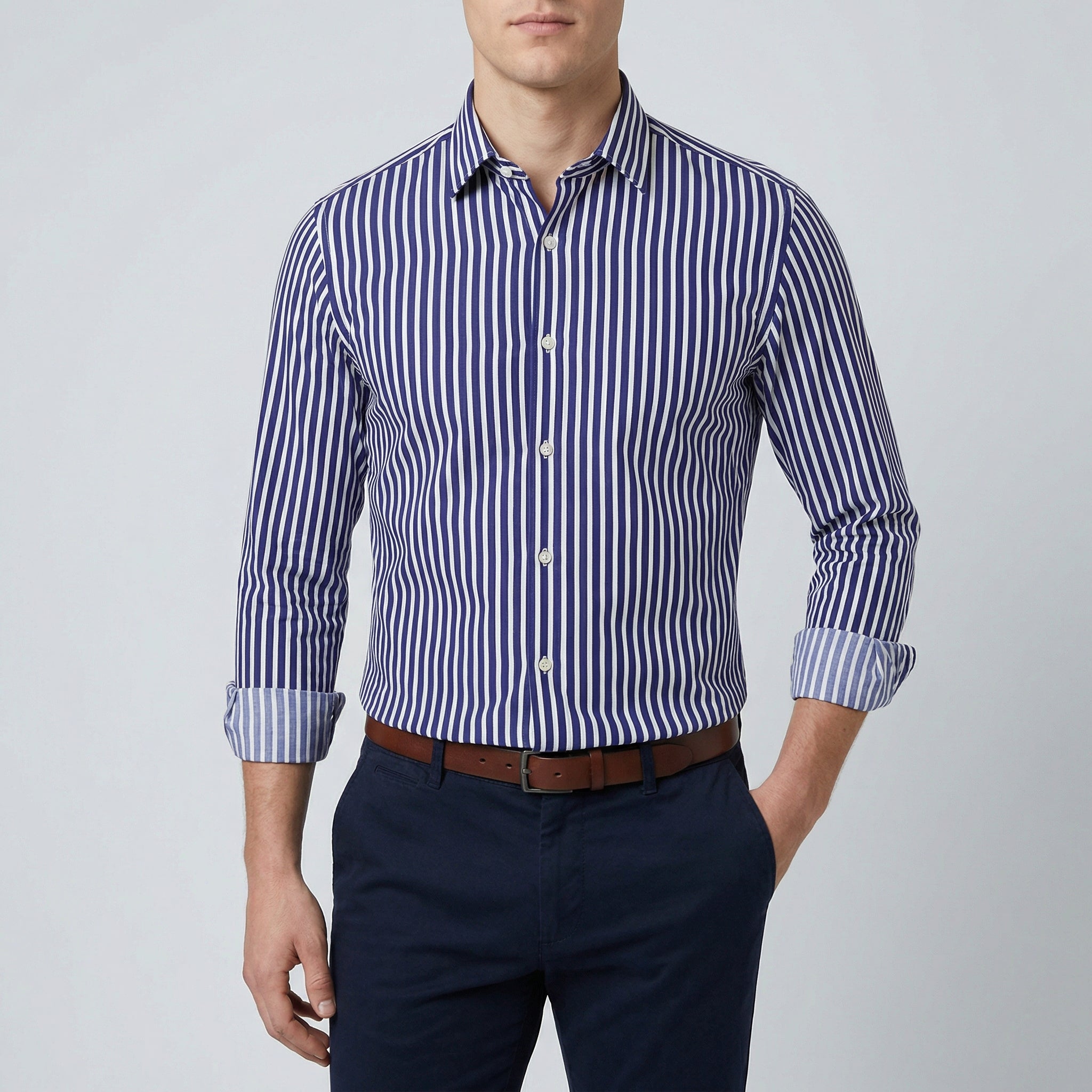 Chemise habillée à rayures bleues et blanches, coupe slim, manches longues retroussées, col boutonné, portée par un homme avec un pantalon marine et une ceinture marron.