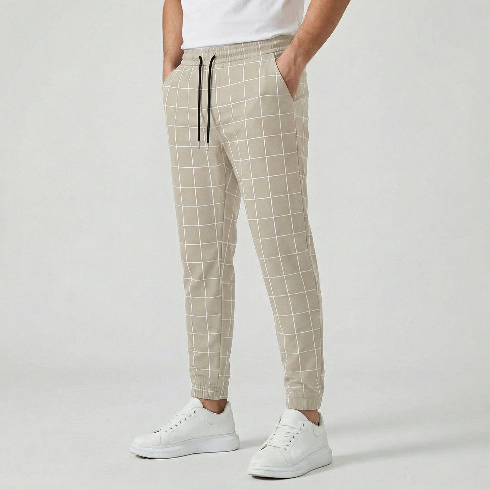 Pantalon jogger à carreaux pour homme, couleur kaki, taille élastiquée avec cordon, poches latérales et chevilles côtelées, porté avec des baskets blanches.