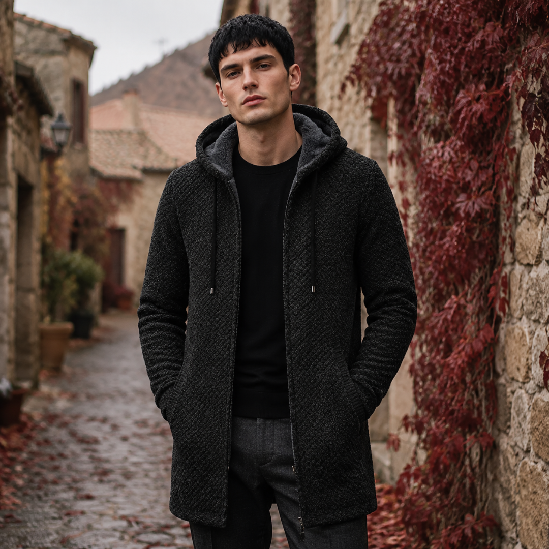 Manteau homme noir à capuche, coupe longue et poches latérales, en pleine rue pavée