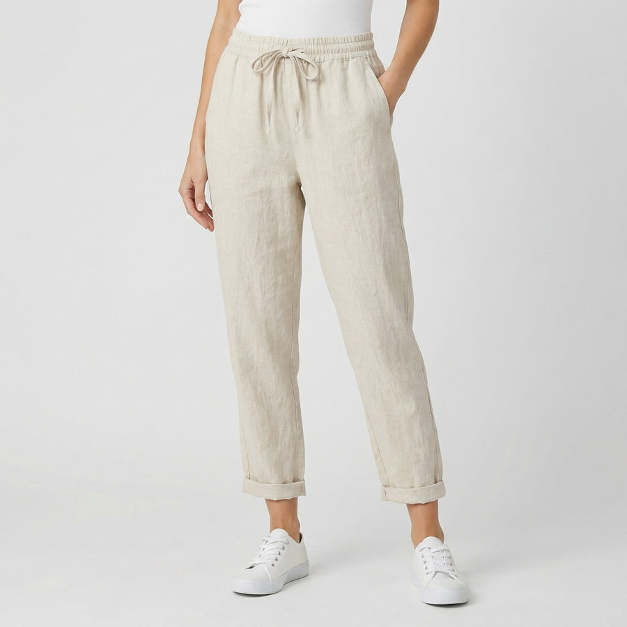 Modèle féminin portant un pantalon jogger en lin beige naturel avec ceinture à cordon, revers aux chevilles et baskets blanches sur fond gris clair