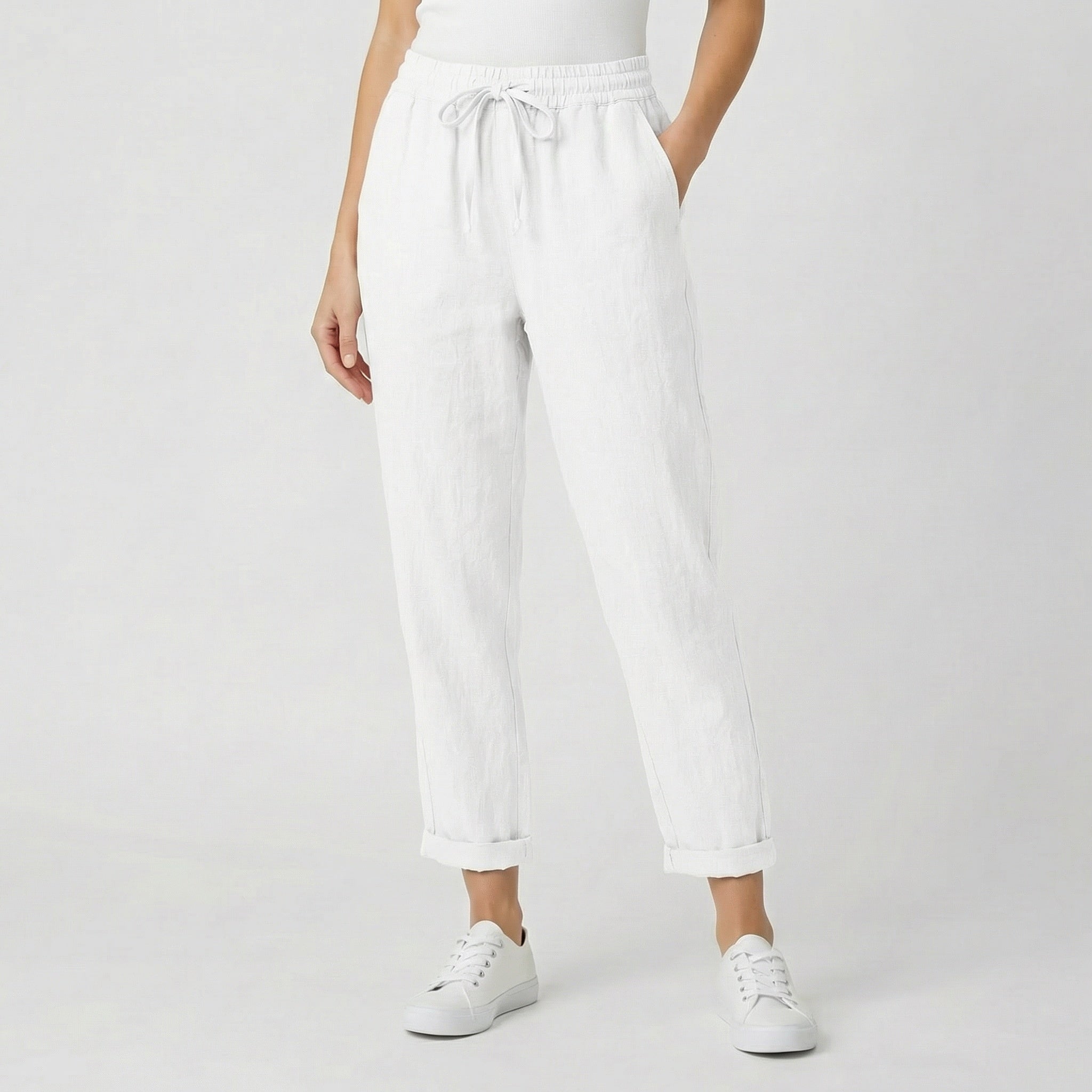 Modèle féminin portant un pantalon jogger en lin blanc avec ceinture à cordon, revers aux chevilles et baskets blanches sur fond gris clair