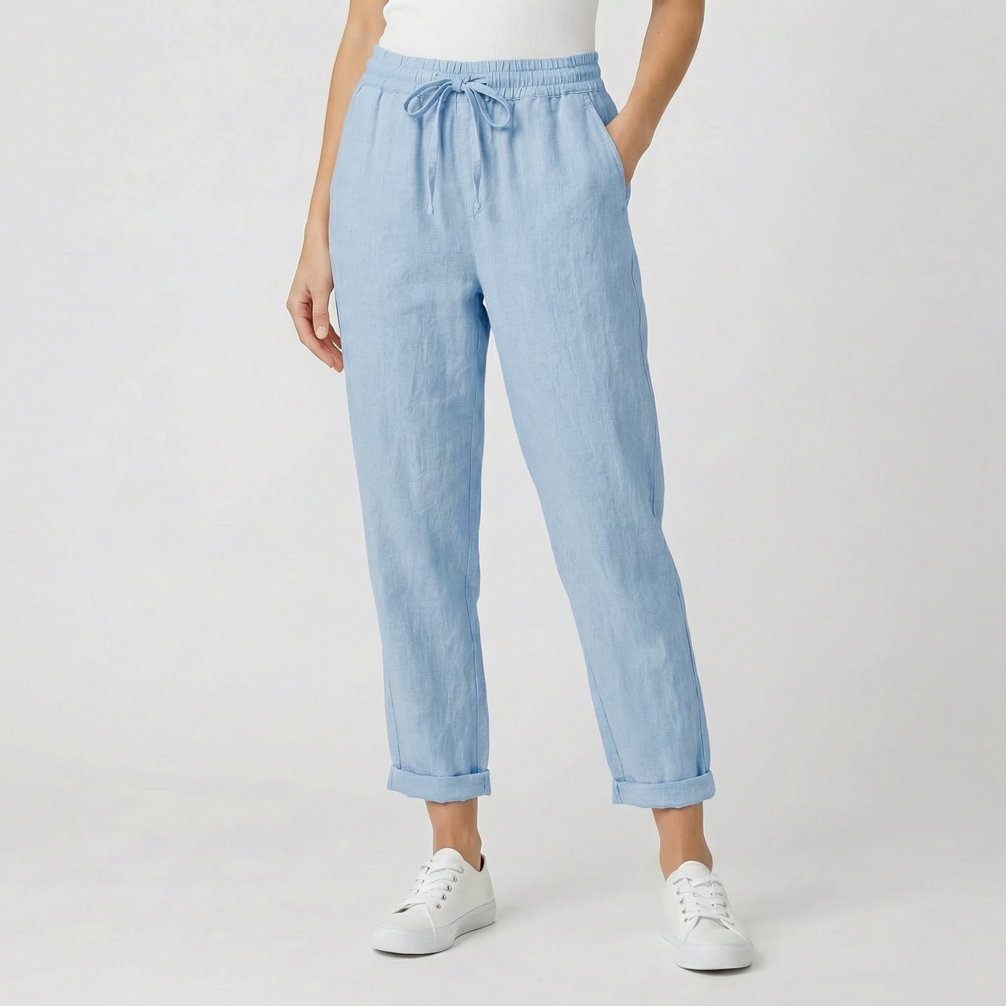 Modèle féminin portant un pantalon jogger en lin bleu clair avec ceinture à cordon, revers aux chevilles et baskets blanches sur fond gris clair
