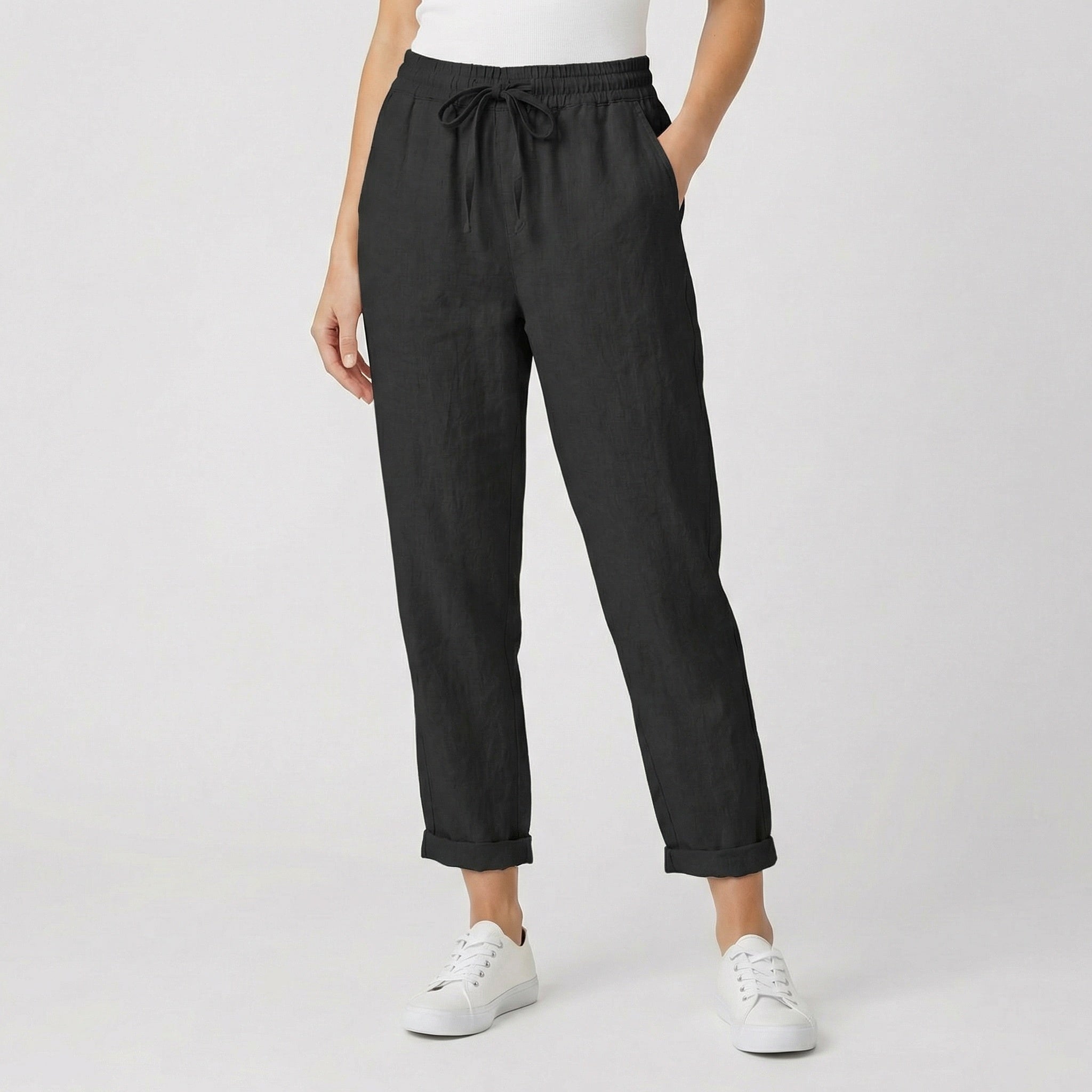 Modèle féminin portant un pantalon jogger en lin noir avec ceinture à cordon, revers aux chevilles et baskets blanches sur fond gris clair