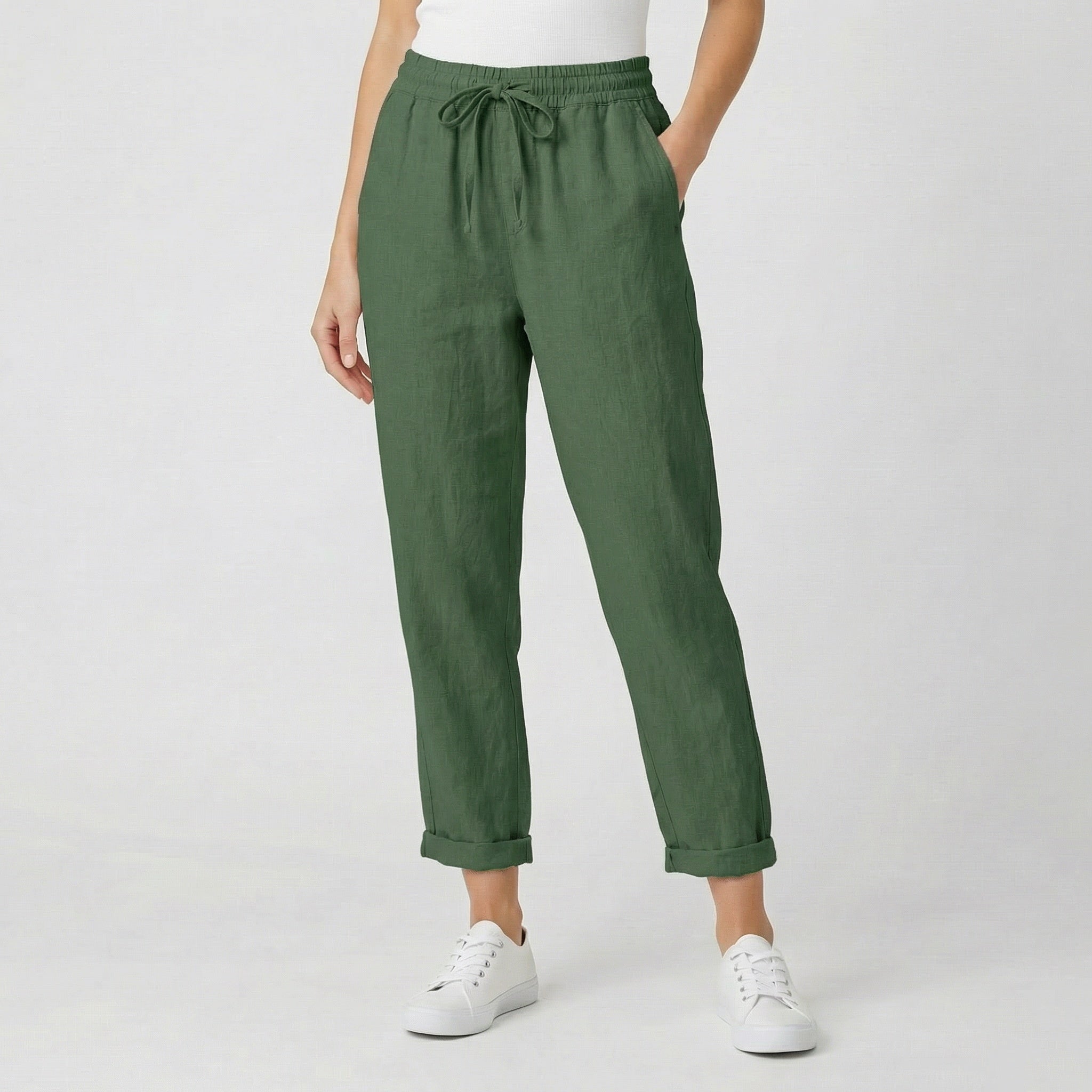 Modèle féminin portant un pantalon jogger en lin vert olive avec ceinture à cordon, revers aux chevilles et baskets blanches sur fond gris clair