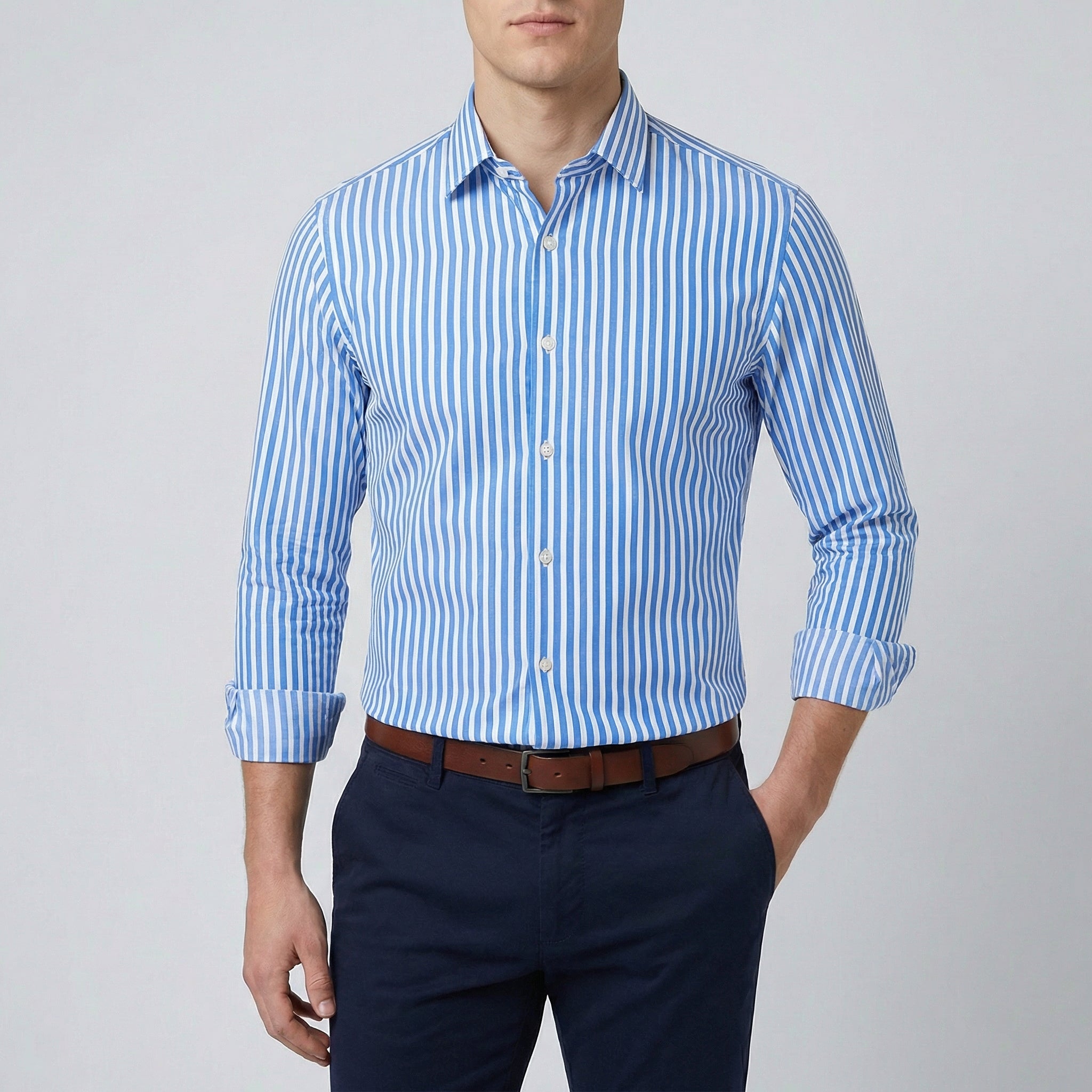 Chemise habillée à rayures bleues claires et blanches, coupe slim, manches longues retroussées, col boutonné, portée par un homme avec un pantalon marine et une ceinture marron.
