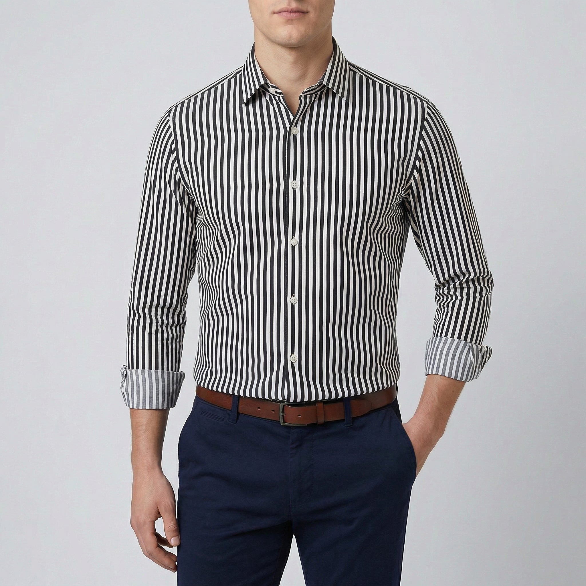 Chemise habillée à rayures noir et blanches, coupe slim, manches longues retroussées, col boutonné, portée par un homme avec un pantalon marine et une ceinture marron.
