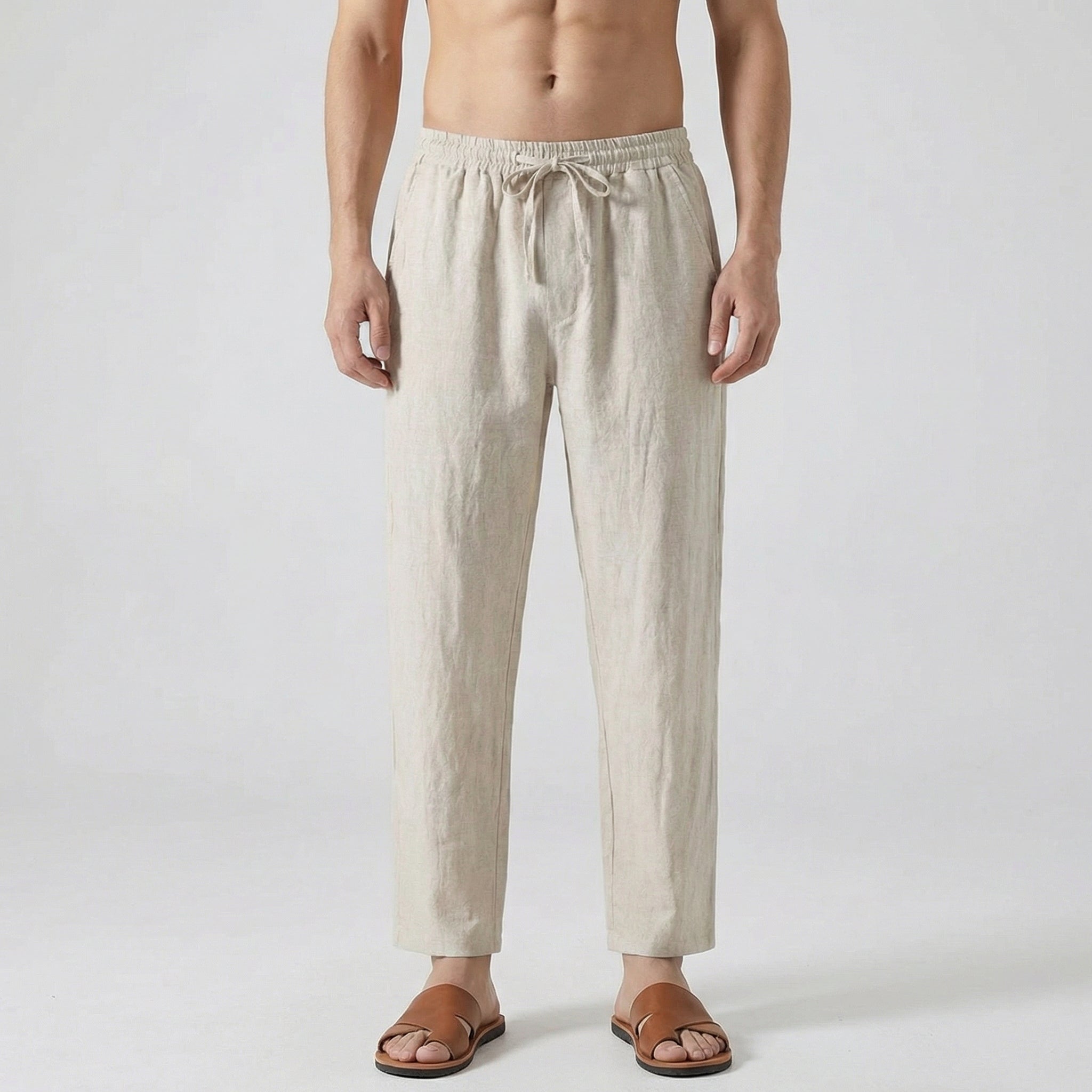 Pantalon en lin beige naturel porté par un homme torse nu, coupe décontractée avec cordon de serrage à la taille, sandales en cuir marron, fond gris clair.
