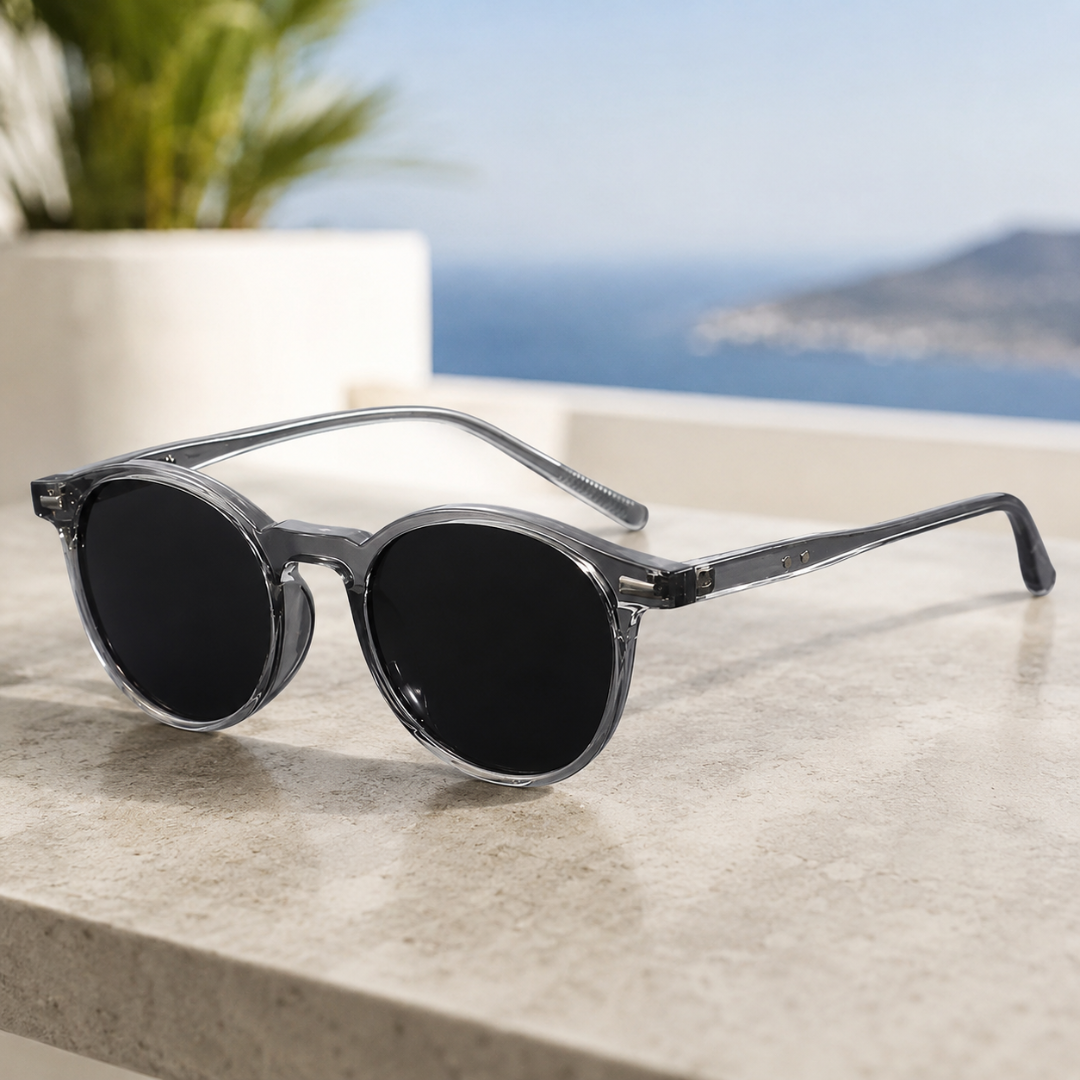 Lunettes de soleil rondes monture grise transparente posées sur un rebord en pierre avec vue sur la mer