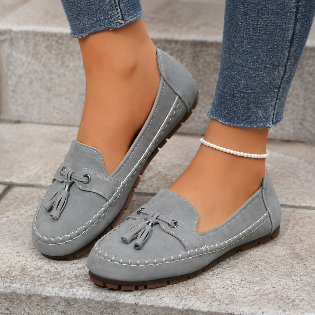 Mocassins plats gris à pompons portés dans un escalier, surpiqûres blanches et semelle crantée